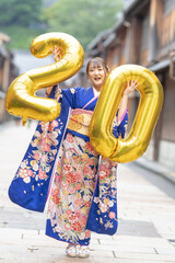 On a summer day, a 20-year-old Japanese girl wearing a blue kimono is in the traditional townscape of a Japanese city. She is holding large golden balloons shaped like the numbers 2 and 0.