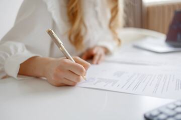 Woman writing on documents at a white desk with a calculator and office supplies in a well-lit...