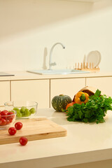 Few cherry tomatoes are scattered on a wooden cutting board, and berries and green grapes are placed in a glass bowl. Vegetables in a paper bag on marble table. Modern kitchen with full equipment.