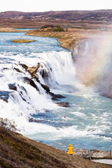 Visitor in yellow jacket admires the powerful Gullfoss waterfall in Iceland, where cascading water meets mist and a vivid rainbow in a dramatic natural landscape.