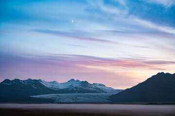 Twilight over Vatnajökull glacier in Iceland, with snow-covered peaks, pastel sky, rising crescent moon, and mist-covered plains evoking stillness and grandeur.