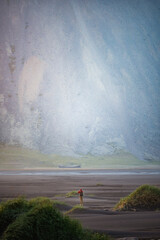 Lone hiker with red jacket walks across black sand dunes in front of the towering cliffs of Vestrahorn mountain in Iceland, evoking solitude and awe in the raw Nordic landscape.