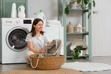 Young woman with wicker basket sitting near modern washing machine in laundry room