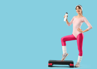 Sporty young woman with water bottle exercising on aerobic stepper against blue background