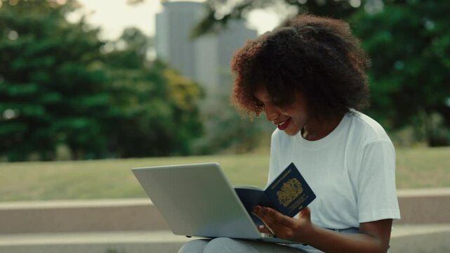 Portrait of cheerful black woman with laptop and passport sitting foutdoors in urban park. Pretty African American female fills visa application online sitting on parapet. Personal data