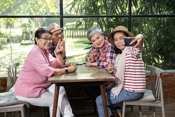 Senior Asian female friends sitting together indoors at modern café making selfie photo smiling flashing peace hand sign sharing joyful casual moment celebrating friendship during cheerful retirement 
