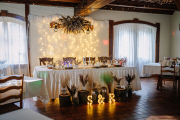 Rustic wedding sweetheart table with fairy lights, dried floral arrangements, candles, and glowing initials “S & R” in a cozy indoor venue..