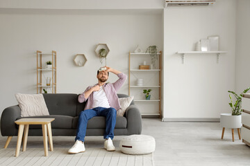 Young man with air conditioner remote control suffering from heat on sofa in living room