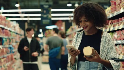 Portrait of smiling African American woman scans a can of coffee with her smartphone standing in supermarket. Female costumer wearing casual clothes checks food contaminants in grocery. Copy space