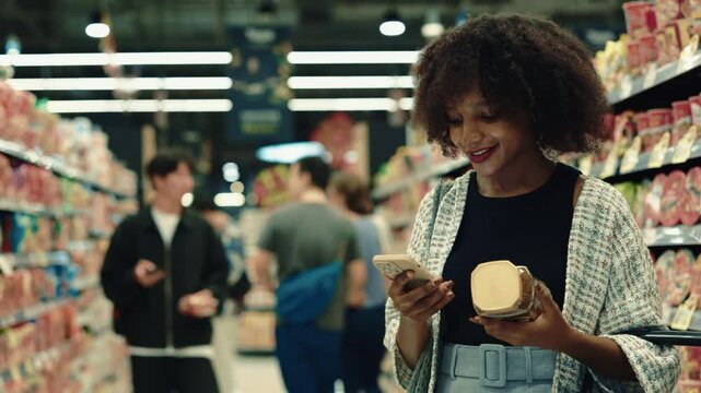 Portrait of smiling African American woman scans a can of coffee with her smartphone standing in supermarket. Female costumer wearing casual clothes checks food contaminants in grocery. Copy space