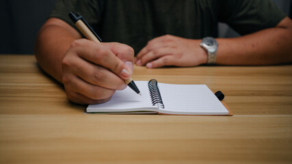 Close-up of hands writing in notebook on wooden desk. Concept of focus, studying, note-taking, or...