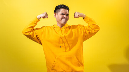 Fototapeta premium Confident young Asian man in yellow hoodie showing biceps with clenched fists and proud expression, standing against solid yellow background. Studio shot symbolizing strength, pride, and energy