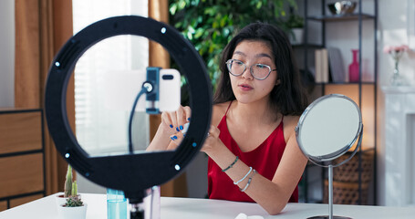Young woman films beauty tutorial with smartphone, gently applying moisturizer. She talks about...