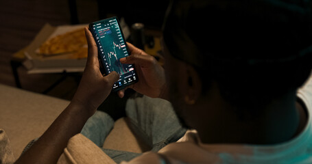 Financial analyst checks real-time cryptocurrency trends on mobile while viewing trading graphs on laptop. Over-the-shoulder angle reveals dual-screen setup.