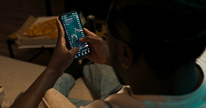 Financial analyst checks real-time cryptocurrency trends on mobile while viewing trading graphs on laptop. Over-the-shoulder angle reveals dual-screen setup.