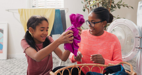 A cheerful girl helps her mom with laundry, both pulling colorful garments from a basket and...