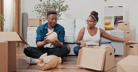 A married couple sits on the floor of their new apartment, unpacking boxes from the move, unrolling...