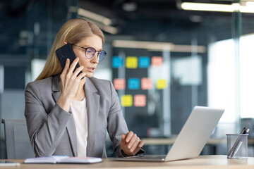 A focused businesswoman in glasses, on a phone call, works on her laptop at her desk. The office has sticky notes on the wall.