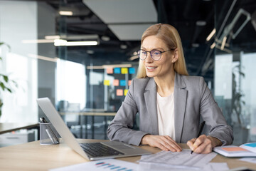 Obraz premium A professional woman in a gray suit works at her desk, focused on paperwork and her laptop in a modern office setting.