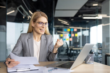 A businesswoman celebrates her success, holding documents and viewing a laptop screen. The office environment suggests a work-related achievement or positive outcome.