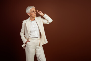 Successful mature businesswoman in stylish white formalwear posing against a brown background with a professional demeanor