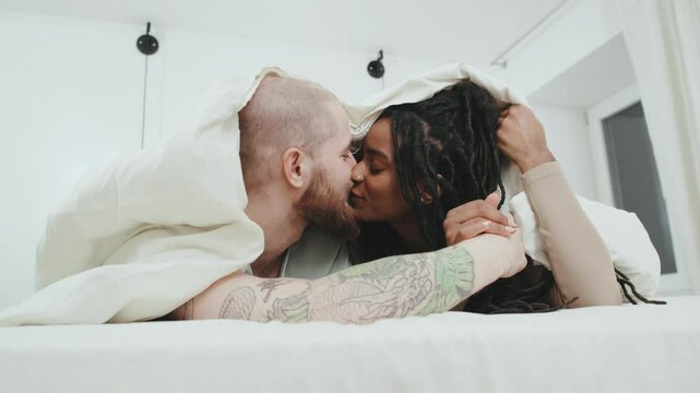 Multiracial Couple kissing while cuddling under a blanket in a cozy bedroom during a warm evening at home