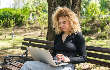 Young university student girl sitting on a park bench, studying and reading material on a laptop...