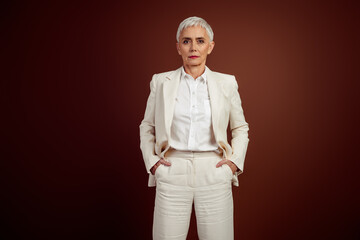 Confident mature businesswoman with short white hair in elegant beige formal suit posing against a brown studio background