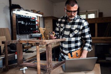 Carpenter standing in his workshop restoring old piece of furniture marking measurements on wood while using a laptop.