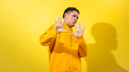 Surprised young Asian man in yellow hoodie stepping back with raised hands and open mouth, looking to the side in fear or shock, posed against a solid yellow background. Dramatic studio shot
