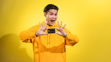Excited young Asian man in yellow hoodie smiling and presenting a credit card with both hands toward the camera against a solid yellow background. Concept of finance, payment, or online transaction