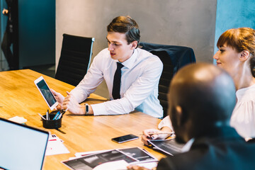 Group of diverse male and female colleagues sitting at conference table with mockup laptop discussing web project during collaborative meeting, multiracial persons teamworking on ideas in company