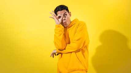 Curious young Asian man in yellow hoodie making an OK sign around one eye like a monocle while looking sideways, posing against solid yellow background. Playful studio concept for exploration