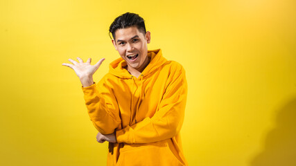 Playful young Asian man in yellow hoodie raising one hand with dramatic sassy expression and open mouth, posing against solid yellow background. Studio portrait for bold, expressive attitude