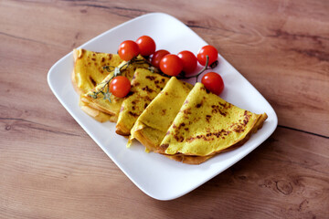 A white plate holds folded yellow crepes garnished with a sprig of vibrant red cherry tomatoes, set on a wooden surface