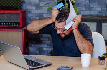 Shocked man with glasses grabbing his head looking at the laptop with a credit card in hand in a...