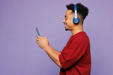 Side view young man of African American ethnicity wear red t-shirt casual clothes listen music in headphones use mobile cell phone isolated on plain pastel purple background studio. Lifestyle concept.