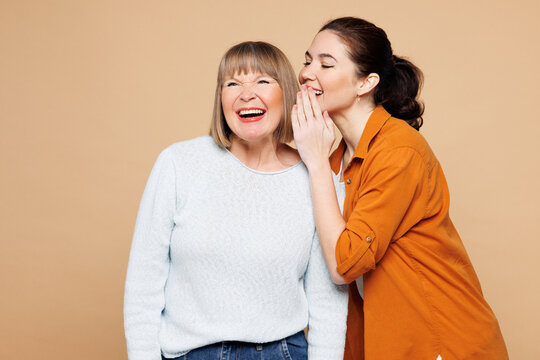 Elder parent mom 50s years old with young adult daughter two women together wears casual clothes whisper gossip and tells secret behind her hand isolated on plain beige background. Family day concept.