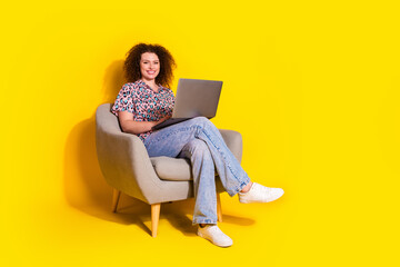 Smiling woman wearing casual outfit using a laptop while sitting on a chair with a vibrant yellow...