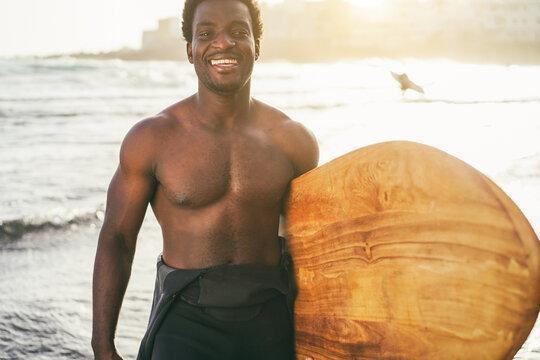 African surfer man holding wood surf board on the beach at sunset - Extreme sport, travel and summer lifestyle concept - Focus on face
