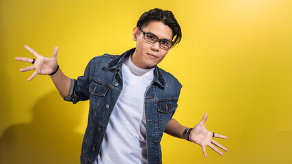 Asian man in glasses and denim jacket striking a fun and expressive pose with arms wide open,...