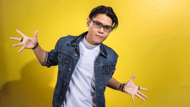 Asian man in glasses and denim jacket striking a fun and expressive pose with arms wide open, smiling slightly, standing confidently against a vibrant yellow background