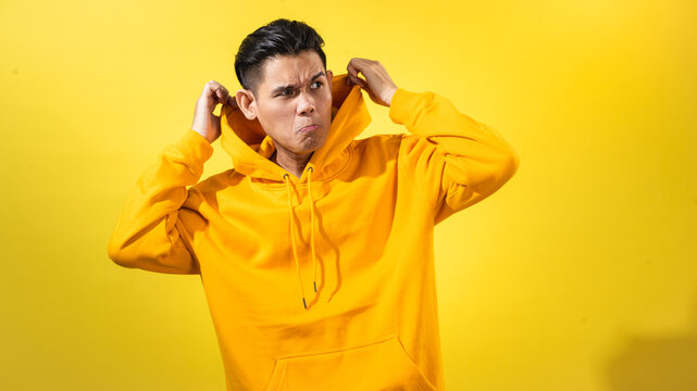 sian man in bright yellow hoodie making a funny expression while pulling hoodie collar with both hands, standing against vibrant yellow background, great for humor, casual fashion, or reaction visuals