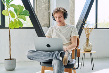 Handsome smiling teenage boy in headphones sitting at home typing on laptop