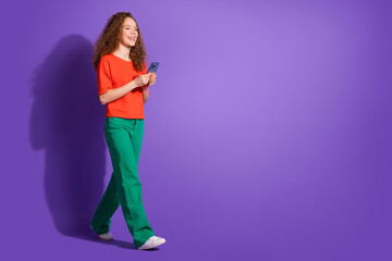 Young woman with curly hair using smartphone while walking against vibrant purple background in casual and stylish orange and green outfit.