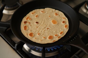 Image of roti / chapatti cooking on cast iron tawa (flat, circular cooking pan), kitchen stove gas burner, wholewheat atta flour, elevated view, focus on foreground