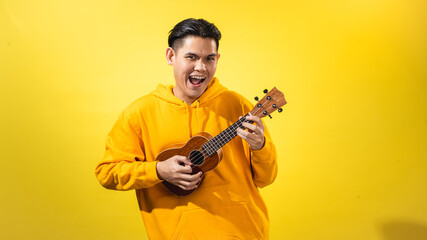 Asian man in yellow hoodie enthusiastically playing a small ukulele against a vivid yellow...
