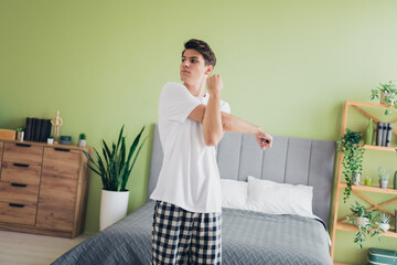 Young man stretching in a cozy bedroom, wearing checkered pajama pants and a white t-shirt, during...