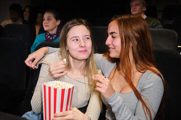 Two friends are enjoying popcorn and whispering to each other while watching a movie at the cinema