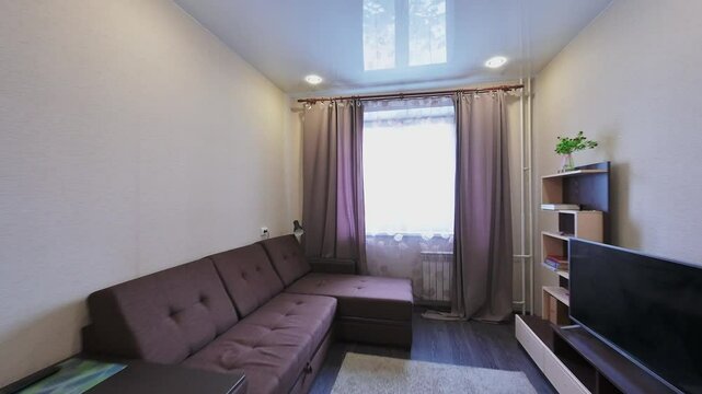 Compact living room features dark brown sectional sofa, minimalist white chair, TV unit, neutral curtains, recessed lighting, and light area rug